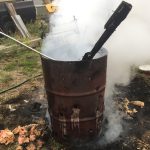 A "burn barrel" in a farmyard is shown smoking and with a piece of unburned wood sticking out. The ground around it is charred.