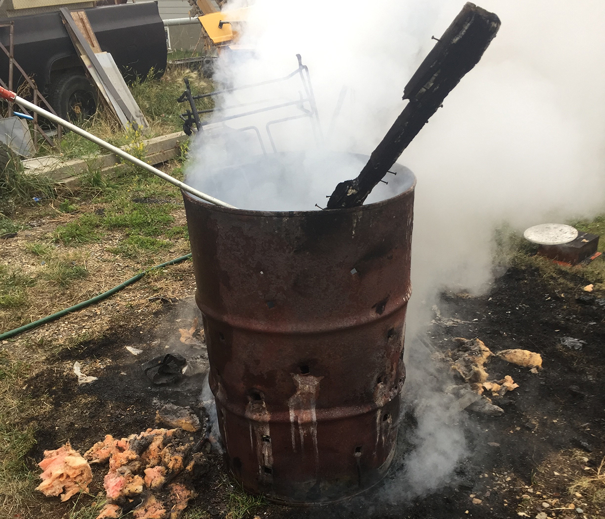 A "burn barrel" in a farmyard is shown smoking and with a piece of unburned wood sticking out. The ground around it is charred.