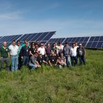 A group of people pose for a photo in front of a large solar panel array in a field.