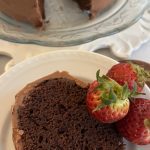 A piece of chocolate cake sits on a plate garnished with fresh, whole strawberries with the whole cake in the background.