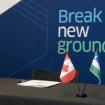 Small flags of both Canada and Uzbekistan sit on a desk with a backdrop that reads "Break New Ground" in the background.