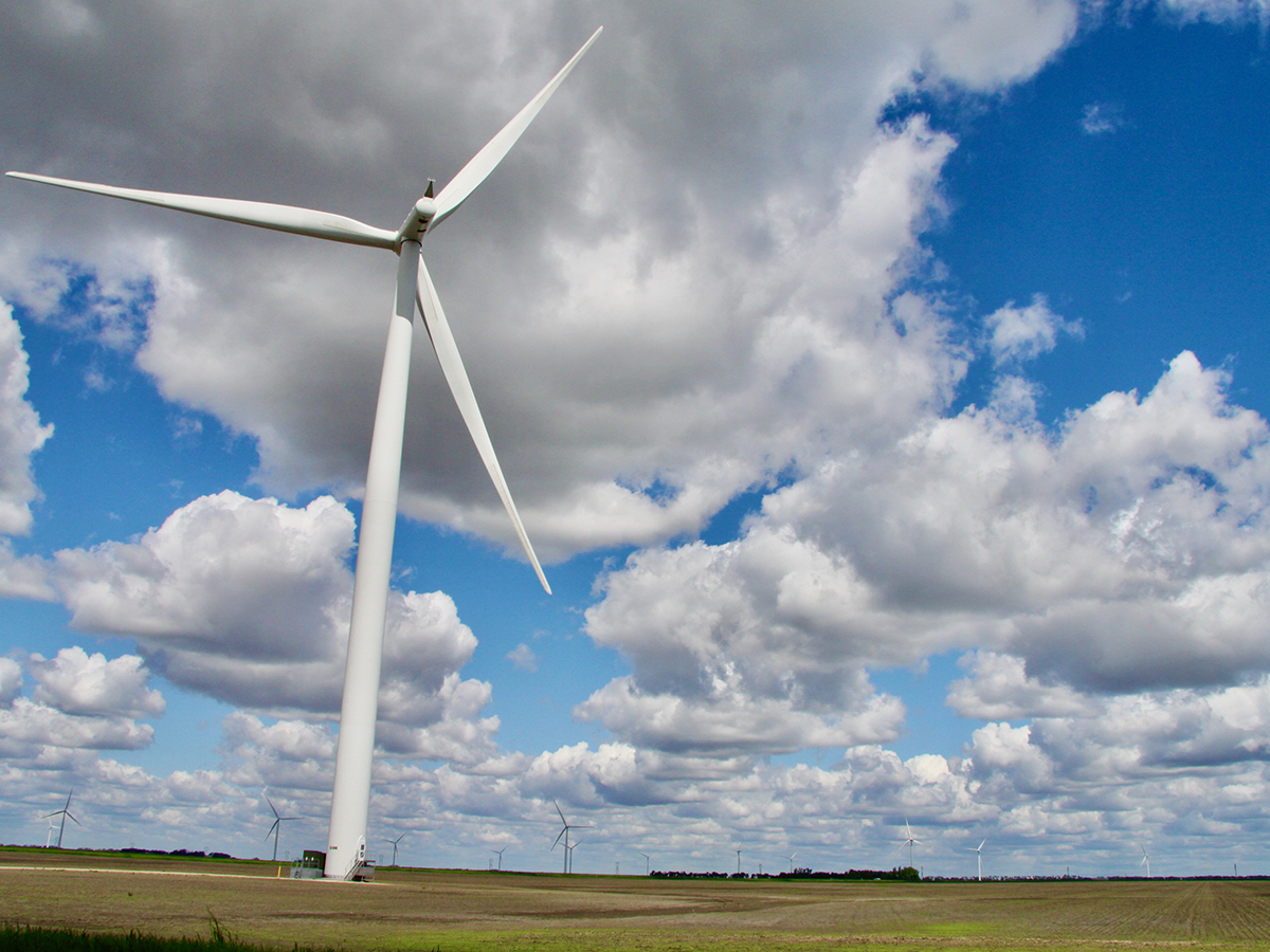 Looking up at a wind turbine on the Prairies against a blue sky with many puffy, white clouds.