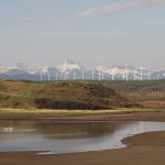 A scenic photo overlooking a small valley with some water and waterfowl in it, a ridge in the background with some wind turbines on it, and the snow-capped Rockies in the background.