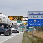 Traffic on a busy road rolls past road signs denoting the upcoming U.S. border, and an LED sign over the roadway showing the approximate length of delay once there.