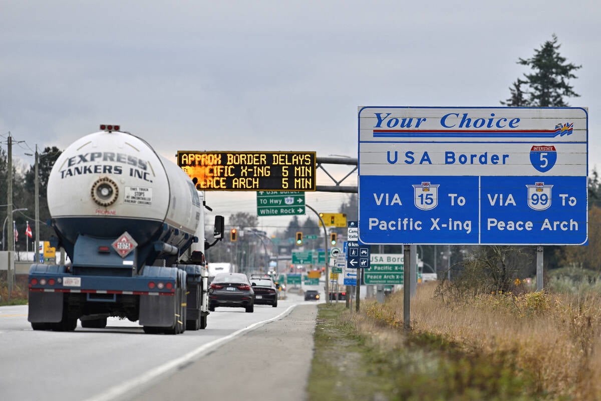 Traffic on a busy road rolls past road signs denoting the upcoming U.S. border, and an LED sign over the roadway showing the approximate length of delay once there.