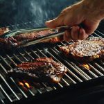 A hand holding a set of tongs flips a steak on a BBQ grill.