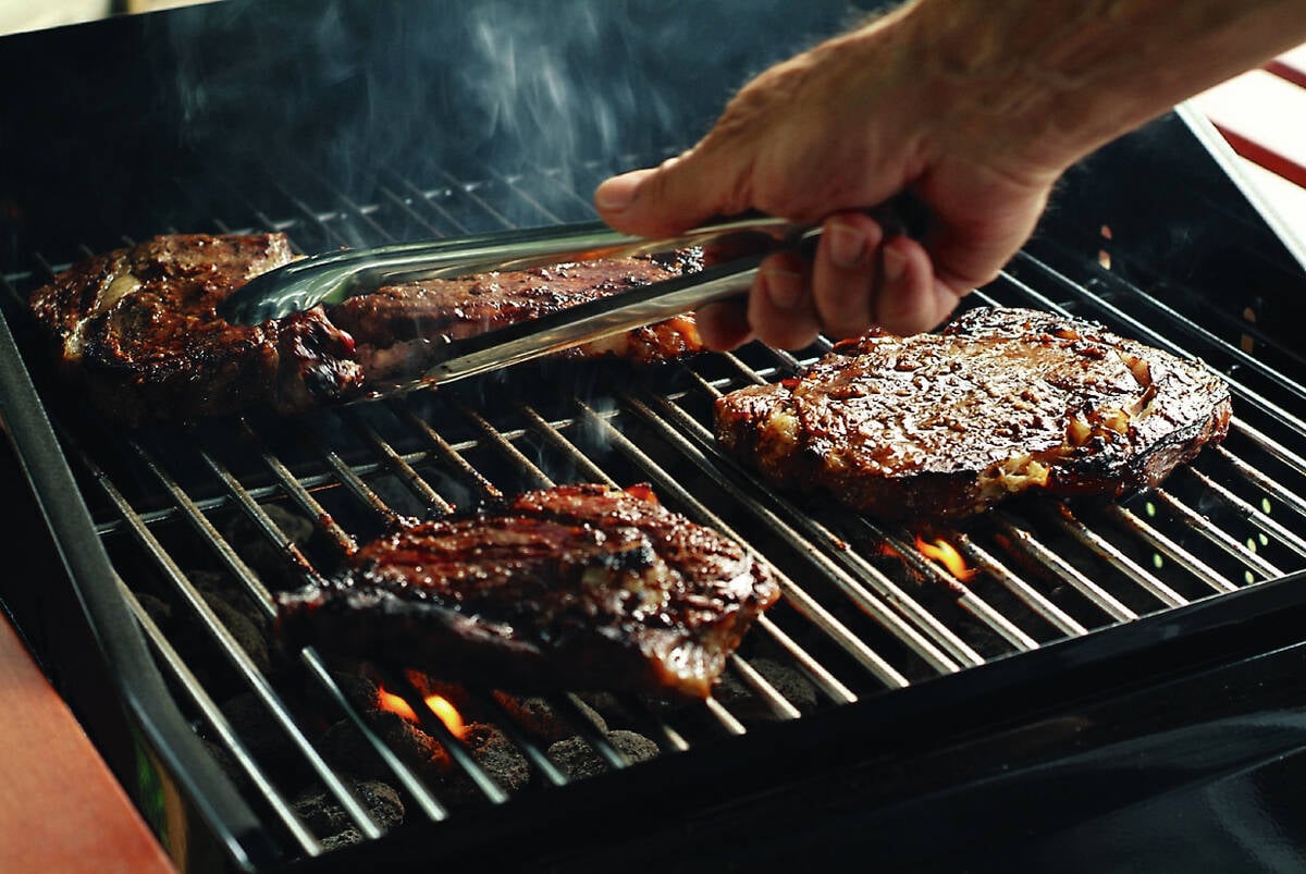 A hand holding a set of tongs flips a steak on a BBQ grill.