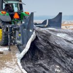 A farmer stands beside a tractor-mounted grain bag roller.
