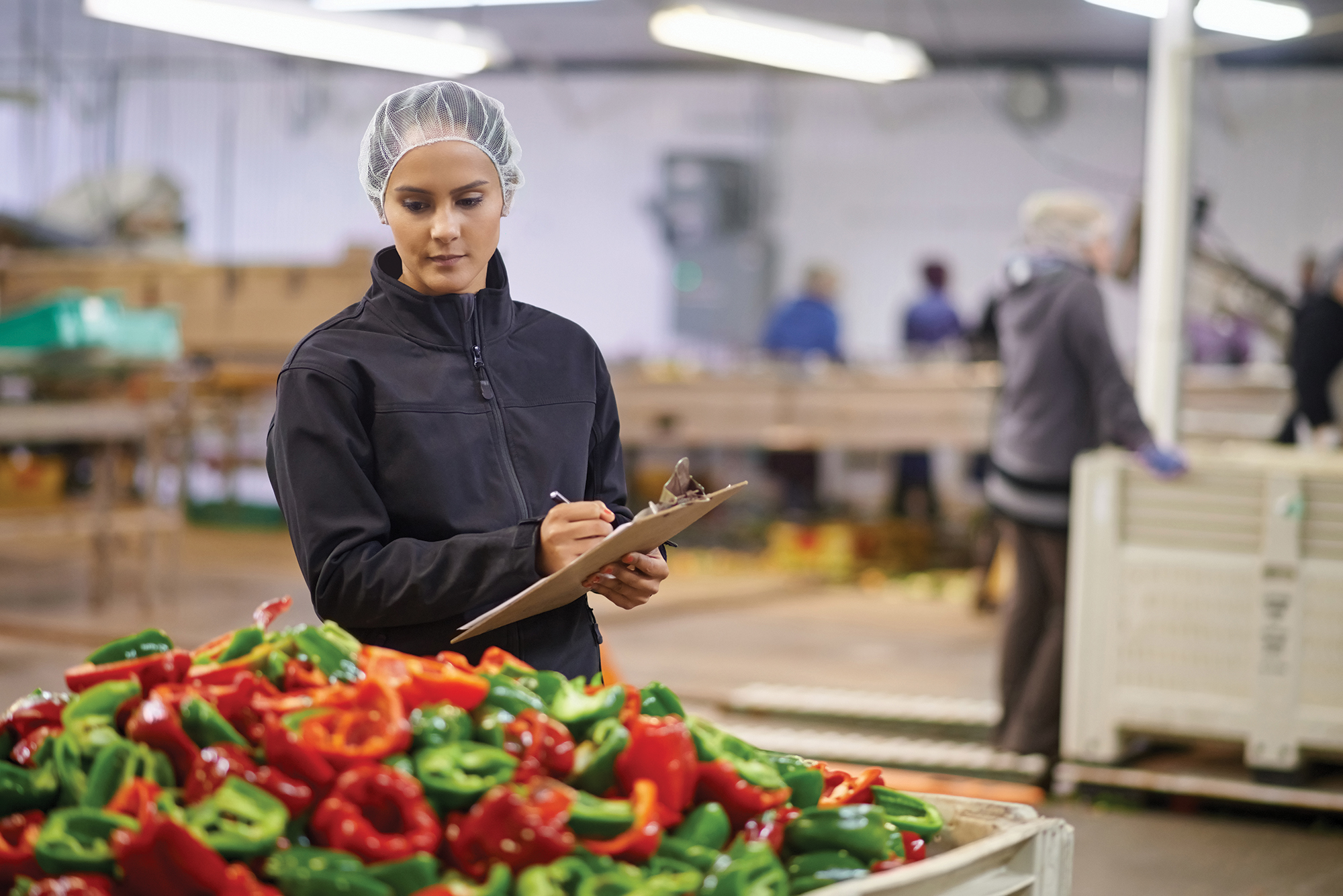 woman in a hairnet and black jacket stands in a food processing facility, writing on a clipboard next to a large bin of chopped red and green bell peppers. Other workers are visible in the background.