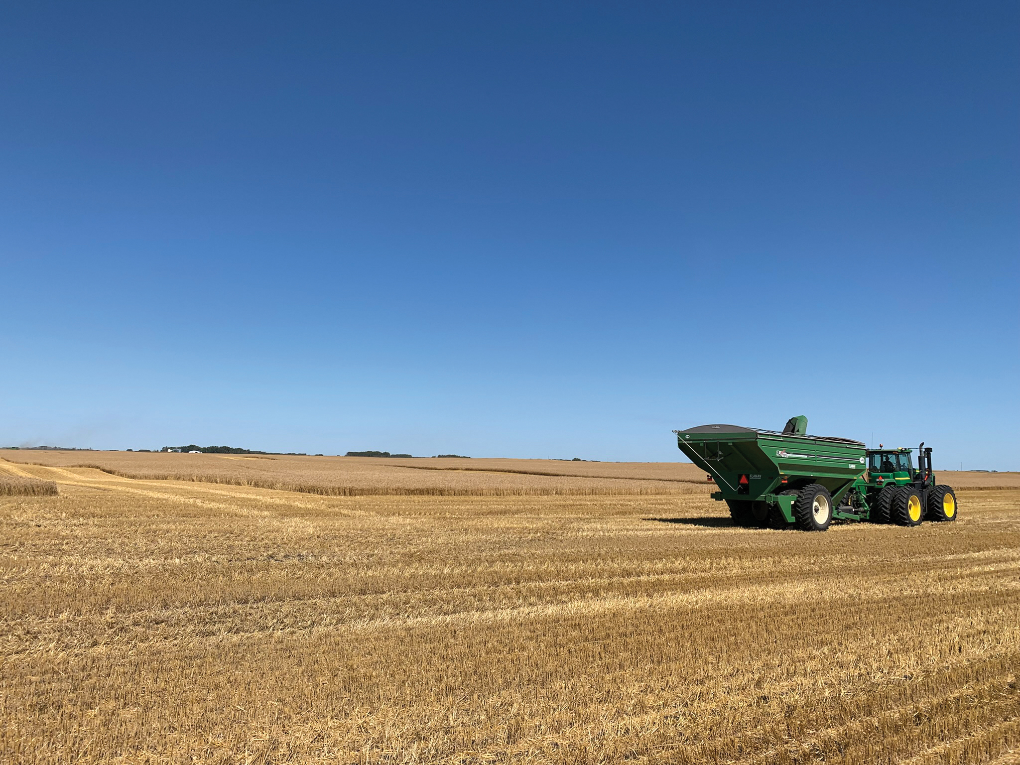 A wide shot of a field with harvested stubble and some standing golden crops under a clear blue sky. A green tractor with a large green grain cart attached sits on the right side of the frame.