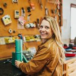 Lesley Kelley smiles for a photo while working with her grain tester in her shop, tools hang on the peg board wall in the background.