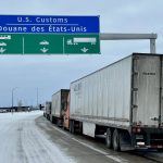 Trucks are lined up on a highway under as sign that reads, "U.S. Customs" as they wait to cross the border.