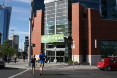 Two people cross a street walking toward a building on a corner with a sign on it that reads, "Future Home of Sunterra."