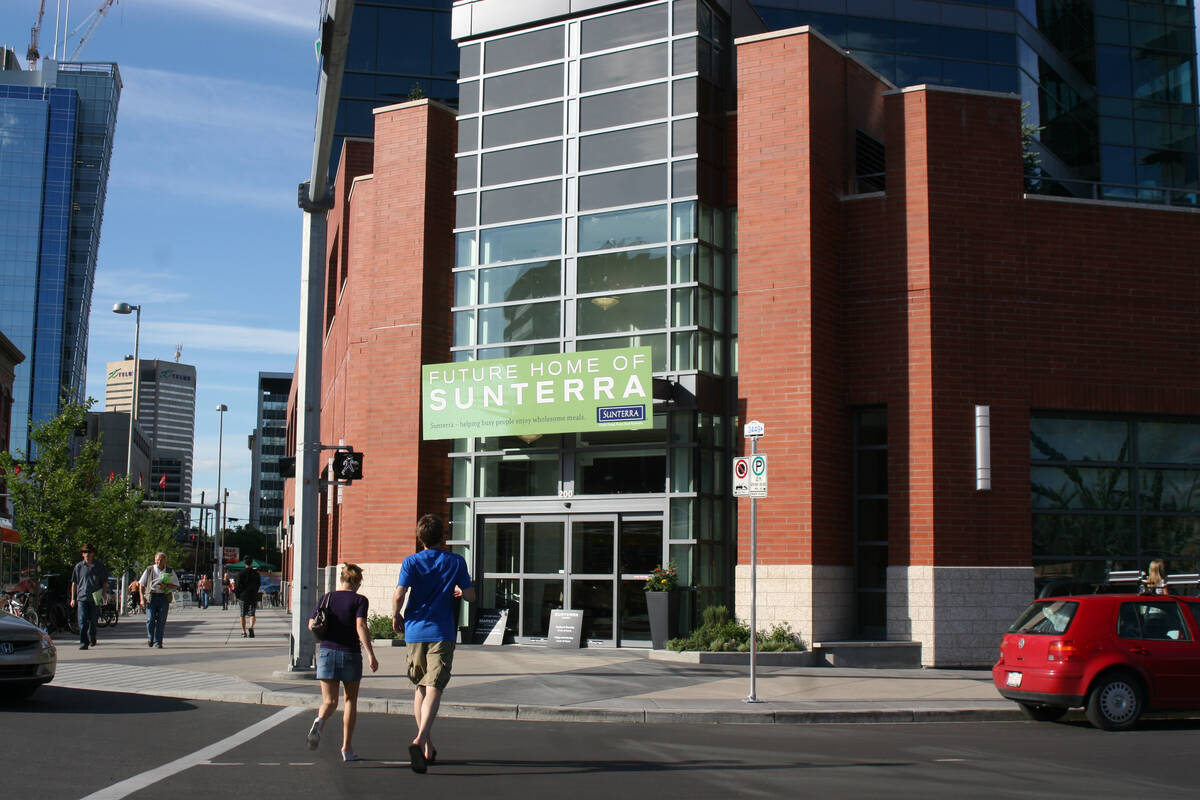 Two people cross a street walking toward a building on a corner with a sign on it that reads, "Future Home of Sunterra."