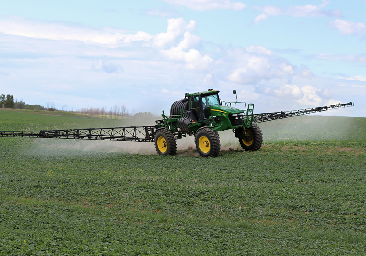 A modern high-clearance sprayer is spraying a crop on a sunny day with large billowing white clouds in the background.