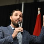 Damien Kurek speaks into a microphone while holding his left index finger up pointing to the sky.