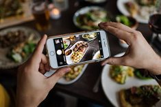 A photo of two hands holding a smartphone taking a photo of a beef dish on a table.
