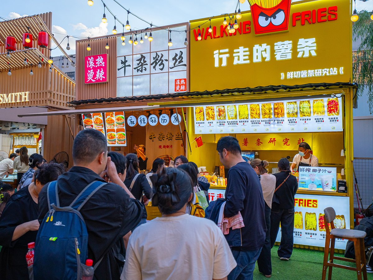 A crowd of people in a Chinese market with food vendor booths in the background, one called, "Walking Fries."