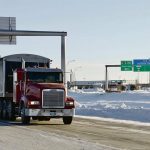 A grain truck is on the highway with a "U.S. Customs" highway sign and border crossing in the background.