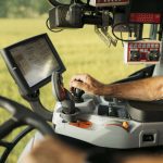 Close-up of older man's right hand on a lever beneath an LCD screen in a modern tractor cab.