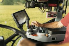 Close-up of older man's right hand on a lever beneath an LCD screen in a modern tractor cab.