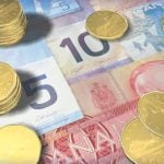 A variety of Canadian currency bills, ranging from $5 to $50, lay flat on a table with several short stacks of loonies on top of them.