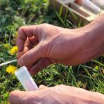 A man's hand is using the small spoon built into the screw-on lid of a plastic tube to collect a small sample of soil in a field.
