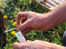 A man's hand is using the small spoon built into the screw-on lid of a plastic tube to collect a small sample of soil in a field.
