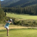 A woman in bare feet tees off on a scenic golf course in the mountains.