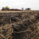 A low angle photo of some disturbed soil with some litter on top of it and an air seeder parked in the background on a cloudy day.