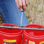 A hand uses a tool to scrape soil from a probe into two red, plastic coffee containers in a field.