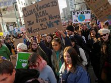 Protesters crowd a street carrying signs that read, "Global warming real. In other news, water is wet," and "Stop denying the [blue painted pic of the Earth] is dying."