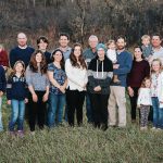 More than 20 members of the Steppler family of the Miami, Man., area pose for a family portrait in some longer grass against a backdrop of trees.