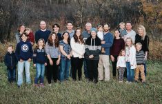 More than 20 members of the Steppler family of the Miami, Man., area pose for a family portrait in some longer grass against a backdrop of trees.