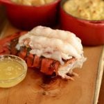 A freshly-cooked lobster tail sits on a wooden table with a small bowl of melted butter beside it.