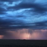 Several strokes of lightning arc downward from an angry dark cloud in the distance near dusk.