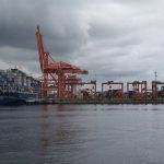 A ship loaded with containers sits at the dock next to several large, orange cranes, at the Port of Vancouver on a cloudy day.