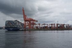 A ship loaded with containers sits at the dock next to several large, orange cranes, at the Port of Vancouver on a cloudy day.