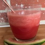 A clear glass with a straw and some red liquid and ice mixture in it sits on a wooden table with some watermelon rind beside it.