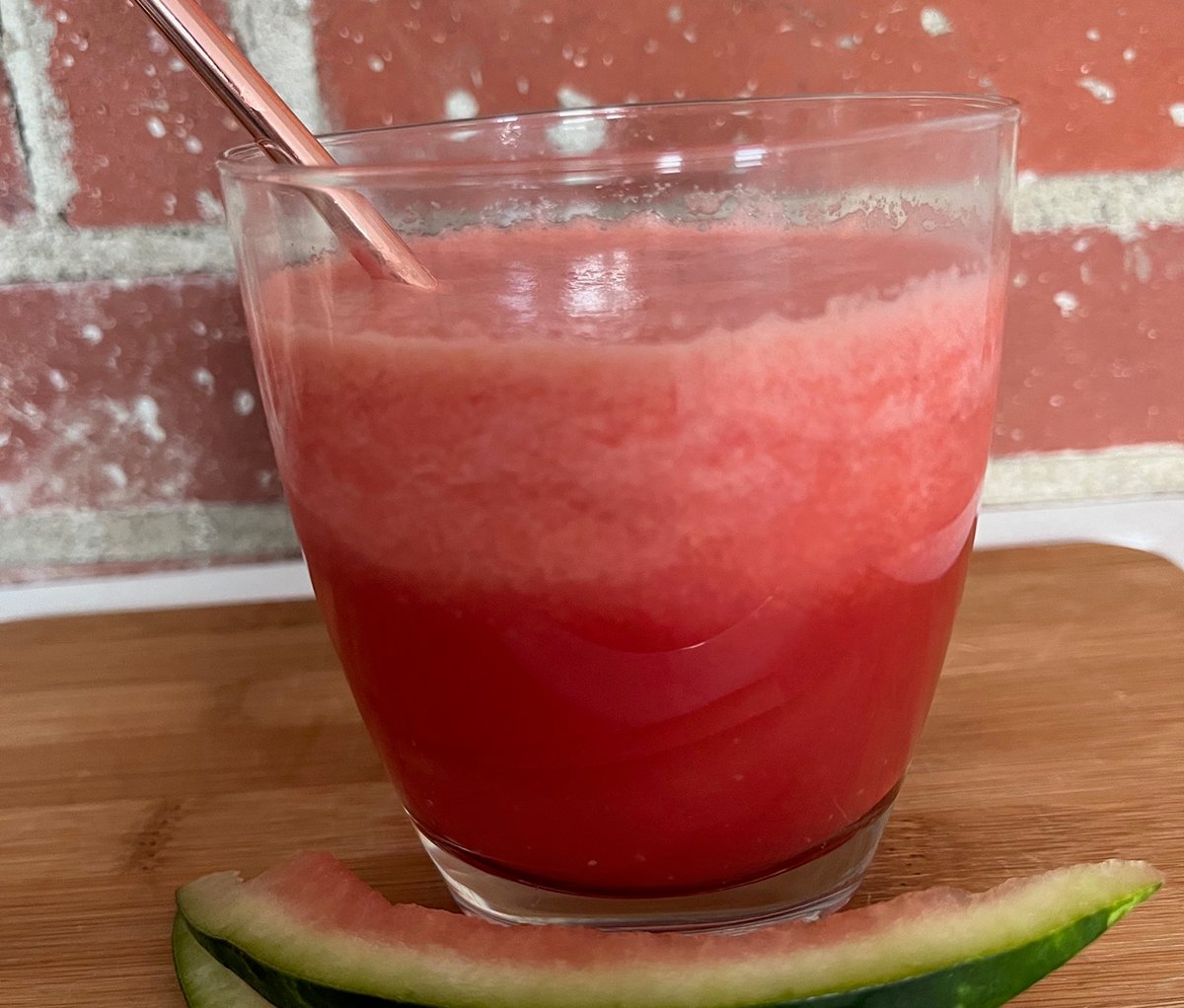 A clear glass with a straw and some red liquid and ice mixture in it sits on a wooden table with some watermelon rind beside it.