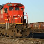 A CN engine sits on the tracks in a rail yard with grain cars visible in the background.
