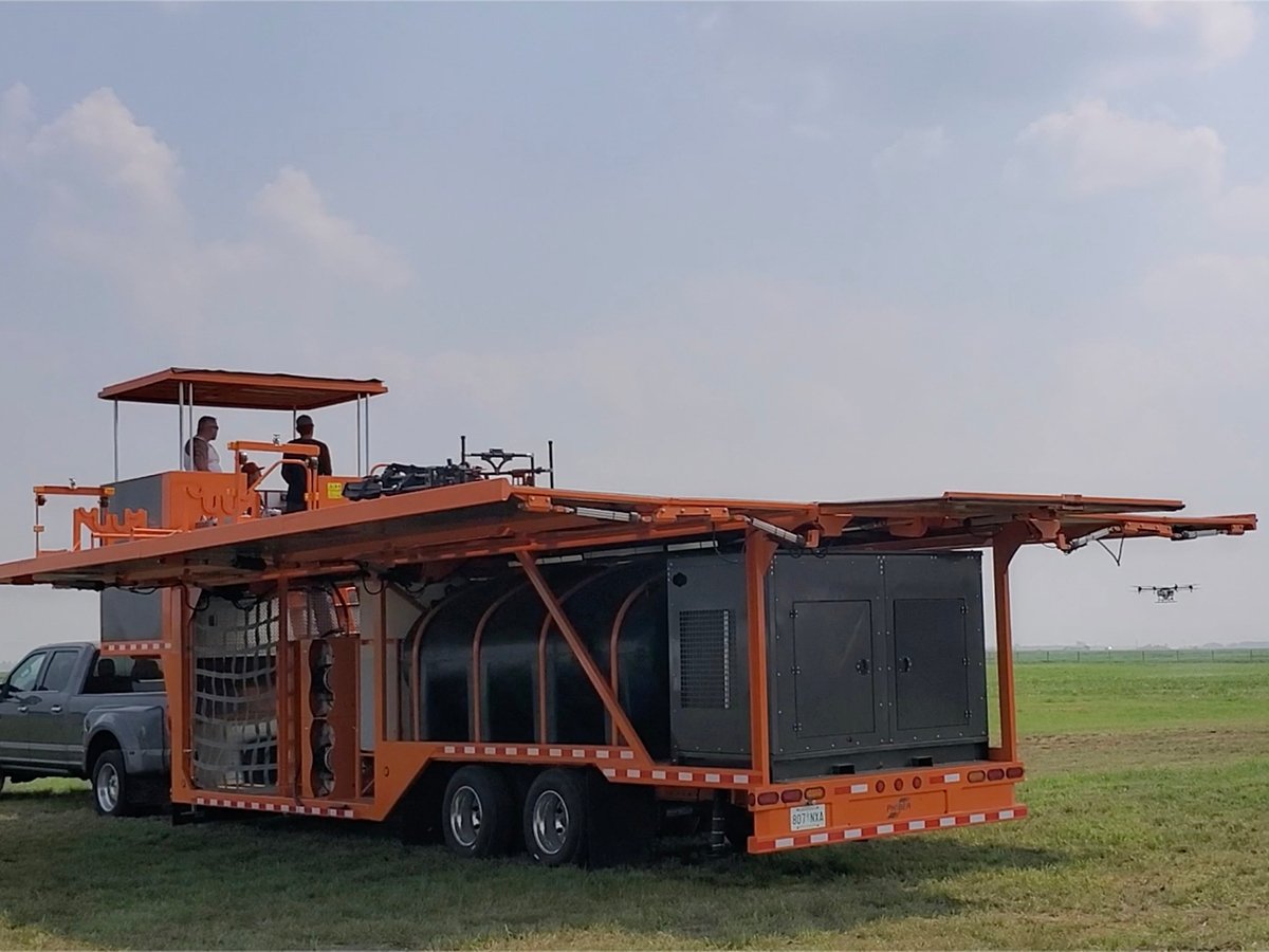 PhiBer Manufacturing's Dash drone tender trailer sits attached to a truck at the drone flying area at the Ag in Motion farm show near Langham, Sask., while operators sit atop the trailer flying a large drone visible in the distance.