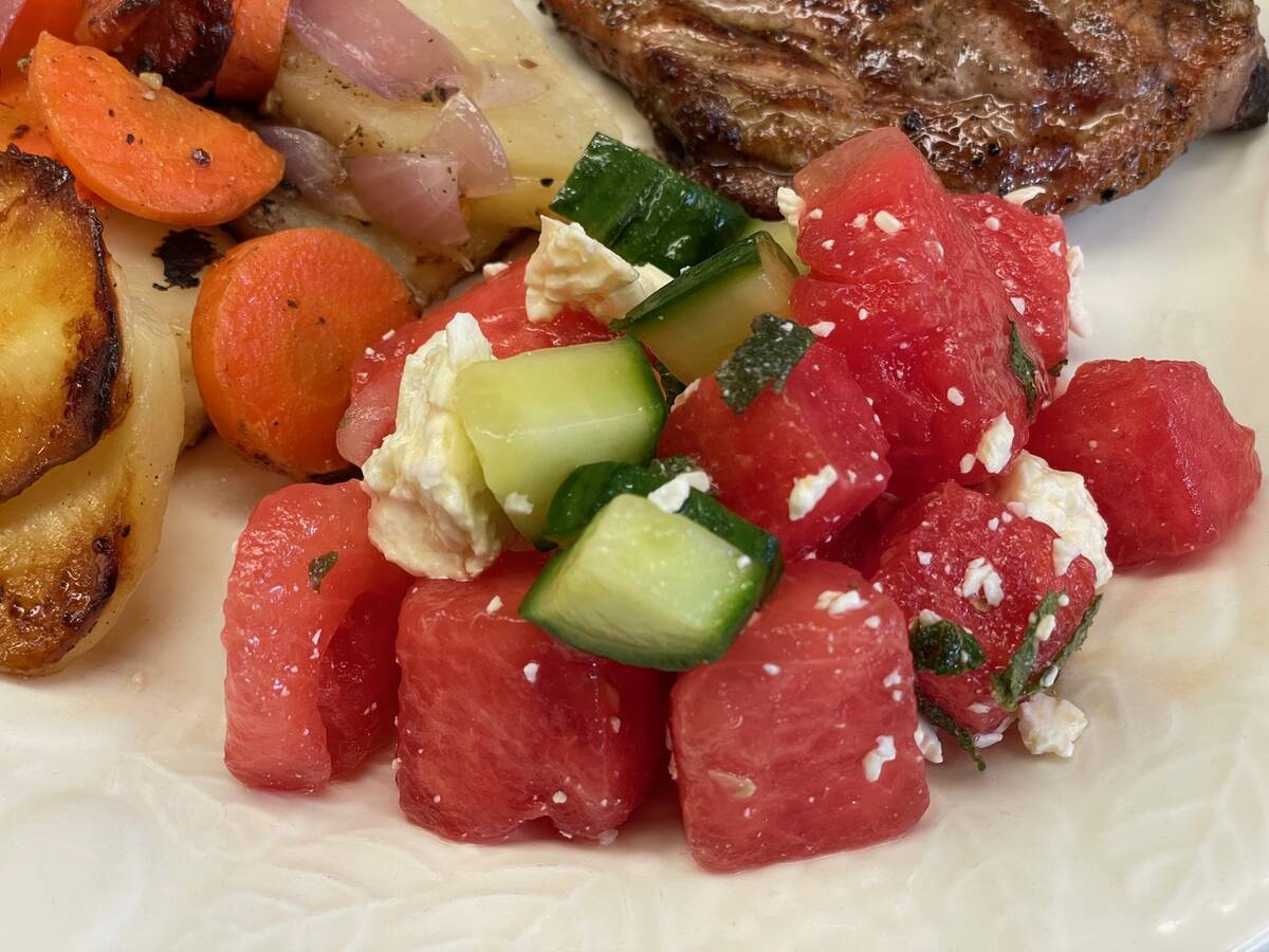 Cubes of ripe watermelon are mixed with chunks of feta cheese, cucumber, red onion and mint on a plate next to a steak and vegetables.