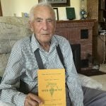 103-year-old Burns Wood, seated in an easy chair and wearing a collared short and suspenders, holds the Alberta Sugar Beet Growers' 50th anniversary celebration booklet from 1975.