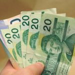 A man's hand holds at least five Canadian $20 bills and two or three $10 bills in the shape of a fan.