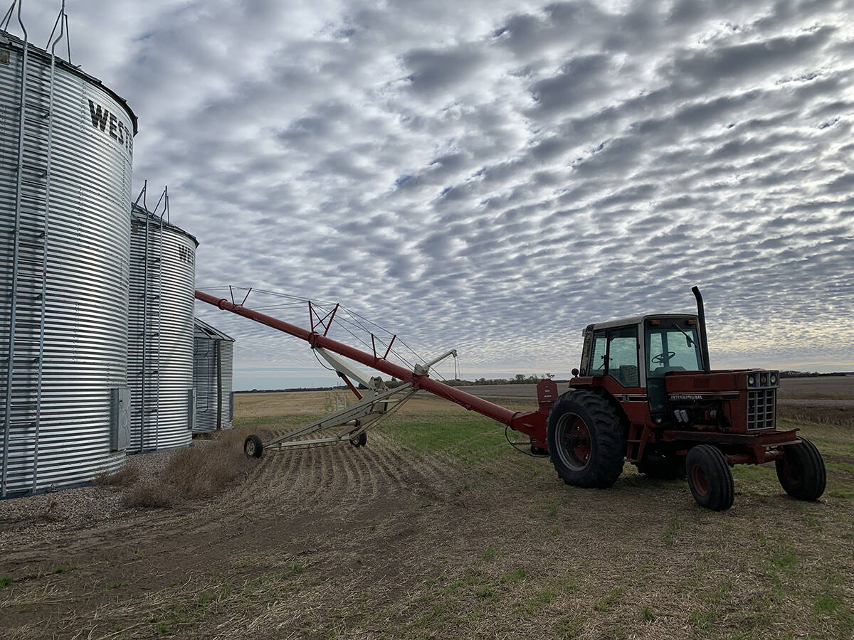 An older tractor with an auger attached to it sits idle next to three steel grain bins under a sky with interesting "rippled" clouds.