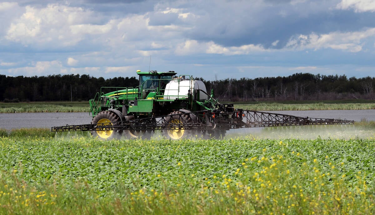 A modern John Deere sprayer sprays a canola crop next to a slough with trees in the background.