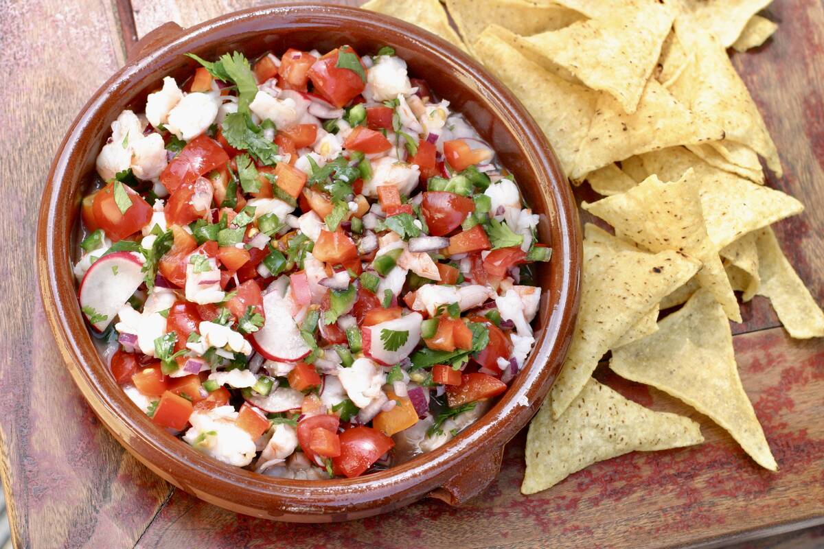 A colourful shrimp ceviche sits in a bowl on a wooden table next to some taco chips.