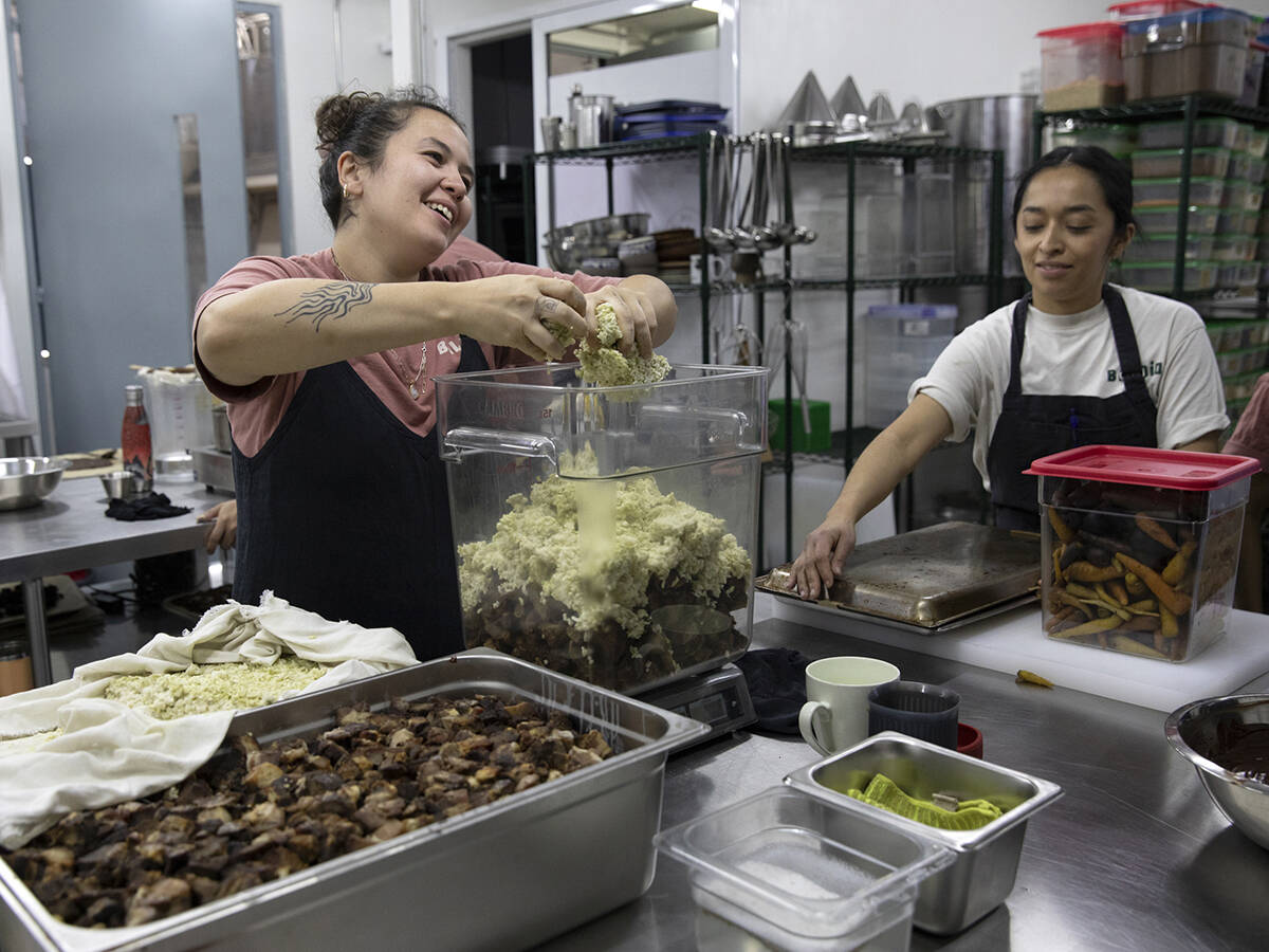 Two women work in a restaurant kitchen, one crumbling rice into a large, clear container with her hands while the other holds a shallow metal pan upside down.