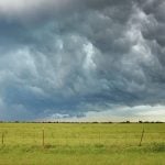 Dark storm clouds over a pasture with a page wire fence in the foreground.